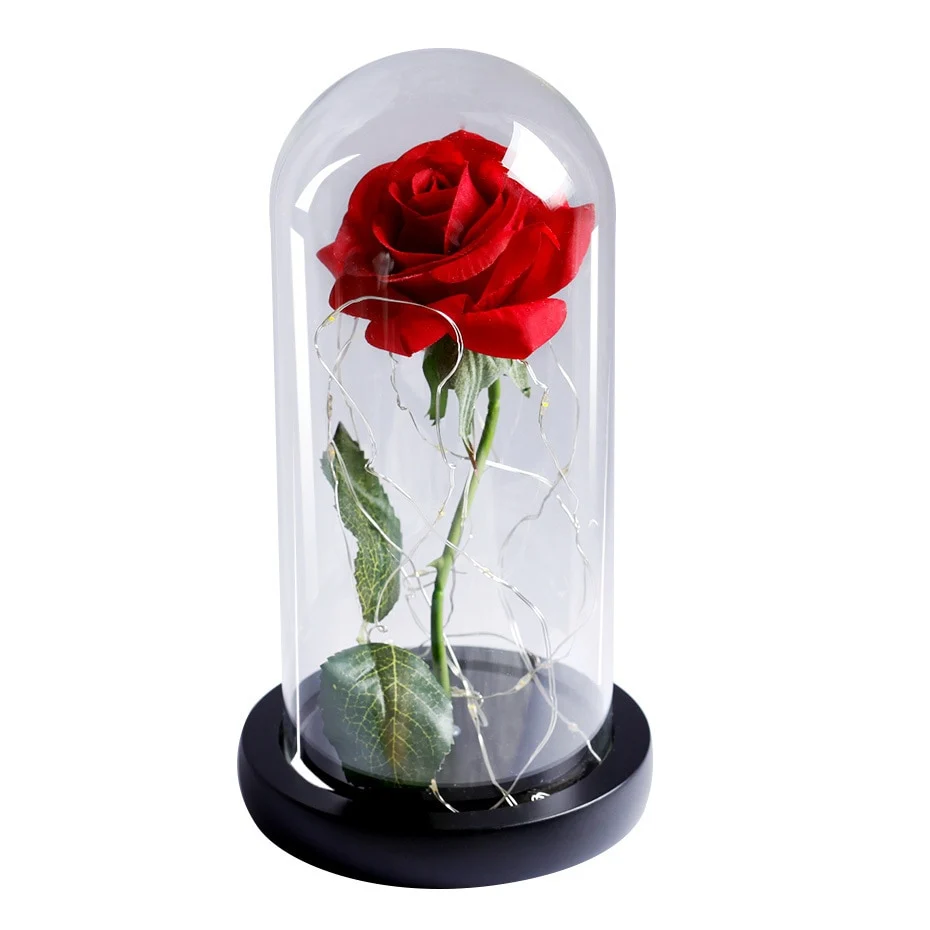 Une rose artificielle rouge sous une cloche en verre avec guirlande lumineuse sur un socle en bois noir, fond blanc